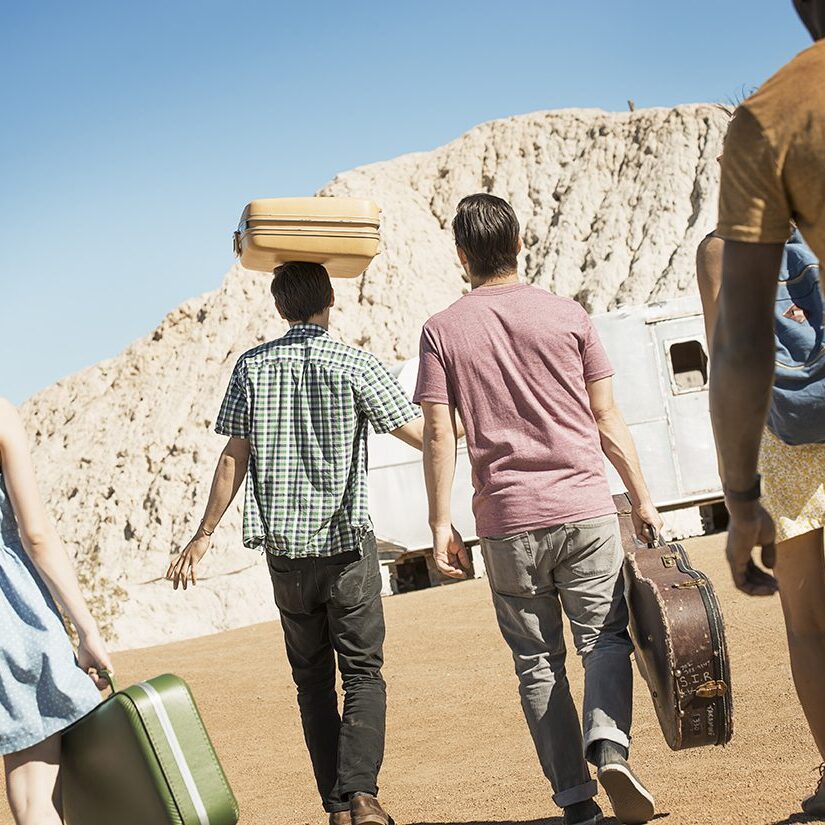 A group of people walking in a line in open desert country, carrying their cases, on a road trip.